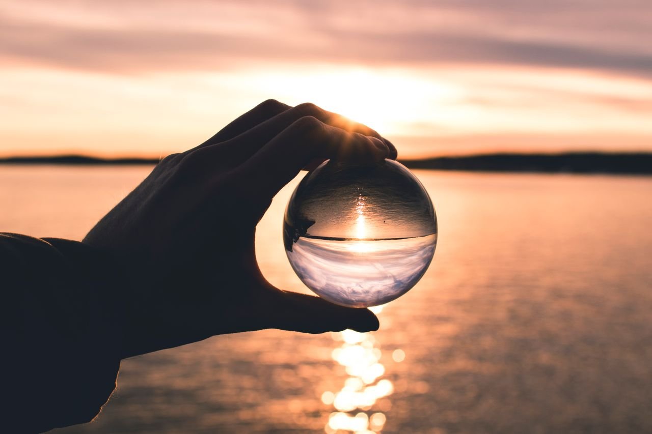 A hand holds a crystal ball reflecting a serene sunset over calm ocean waters.